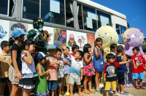 ラッピングバスを背景に琉神マブヤーと記念撮影する子どもたち＝２７日、県奄美パーク
