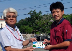 平安町長（左）から知名町ご当地ナンバープレートの交付を受ける田原さん＝１日、知名町役場前