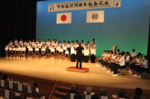 知名中学校吹奏楽部の演奏で、知名町町歌を斉唱する計７０人のフローラル合唱団＝６日、知名町おきえらぶ文化ホールあしびの郷・ちな