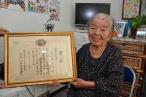 「母から子への手紙」コンテストで１５周年記念賞を受賞した佐々木キヨさん。後ろに見えるのが貞進さんの写真＝２２日、和泊町