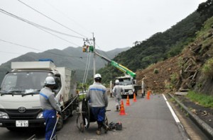 写真ヨコ 奄美市の市道・山間―戸玉間の土砂崩れ現場で行われた高圧配電線などの復旧作業＝７日午前１１時半ごろ、同市住用町