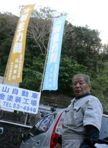 特注のぼりのもとで笑顔を見せる安山さん＝１６日、奄美市名瀬鳩浜町