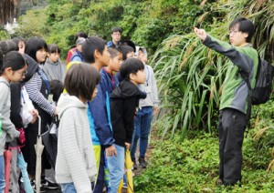 牧野さん（右）の外来種アメリカハマグルマ(下)の説明に聞き入る受講生ら＝７日、瀬戸内町