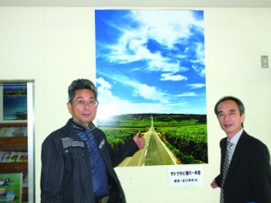 「サトウキビ畑の一本道」パネル展示が始まった支庁喜界事務所。左は撮影した吉行さん＝１４日、喜界町赤連